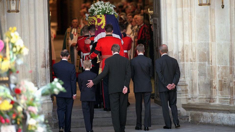 five men in suits walk behind a coffin covered in a multicolored flag and flowers as the procession enters a stone building