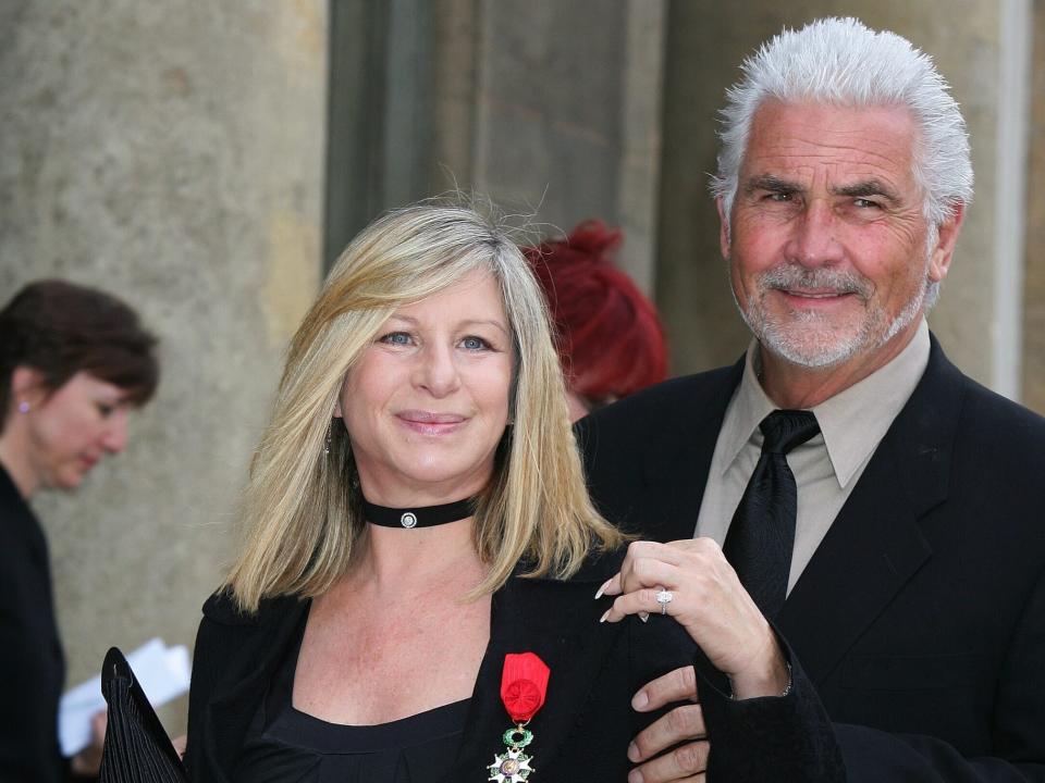 Barbra Streisand, in a black shirt and blazer, stands with a medal pinned to her lapel. Husband James Brolin, in a black suit, stands behind her with his arms around her.