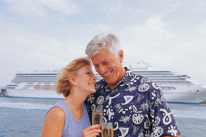 A couple smiling while sipping champagne in front of a docked cruise ship.