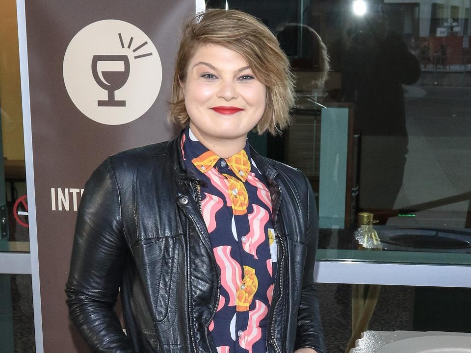 Kylie Sparks wearing a black leather jacket and a bright shirt while standing in front of a window.