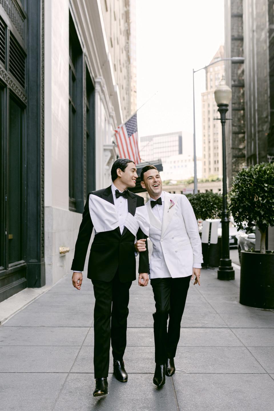 Two grooms look at each other and smile on a busy street.