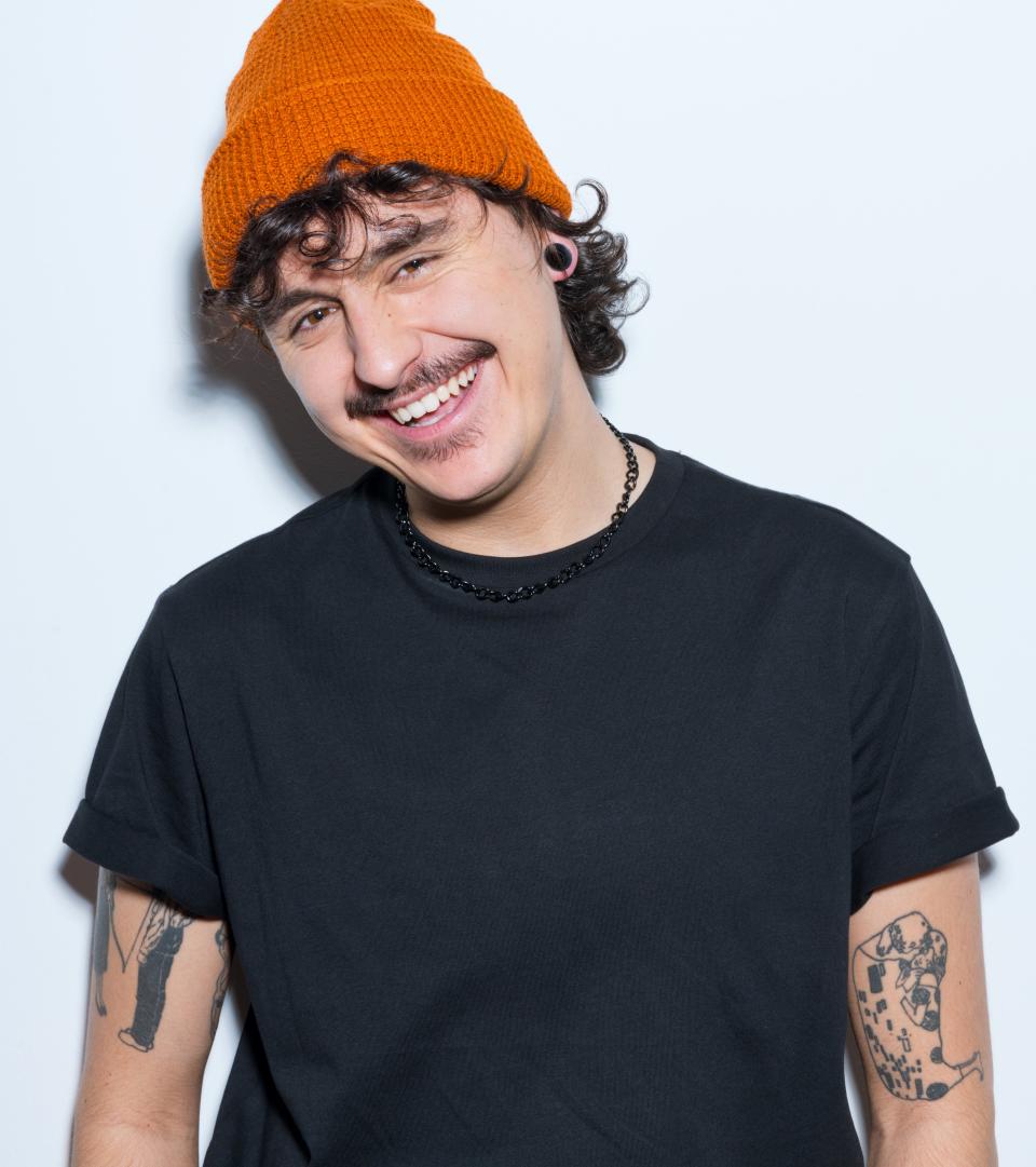 A person with curly hair wearing an orange beanie and a black T-shirt smiles at the camera. Tattoos are visible on their arms