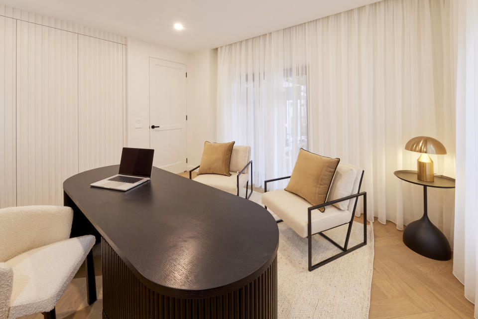 A large lack desk in the centre with three chairs around it, white flowing curtains on the far wall. 