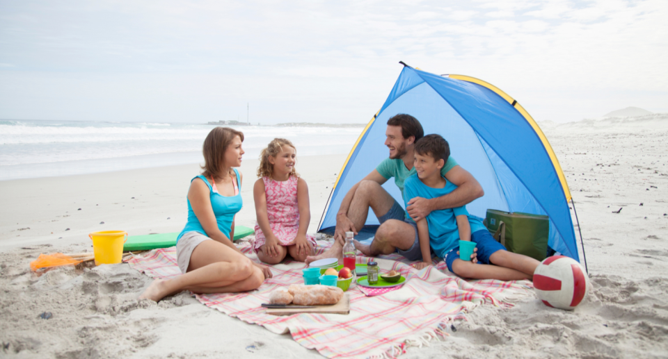 mom and dad with two children sitting on beach blanket with blue beach tend and lunch and snacks