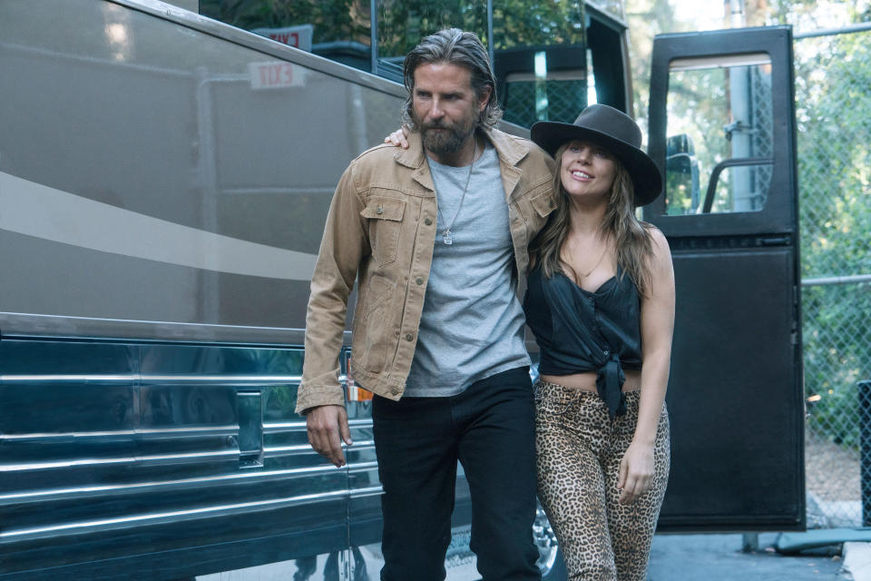 Bradley Cooper in a beige jacket and Lady Gaga in a hat and leopard print pants stand closely together, smiling, beside a tour bus