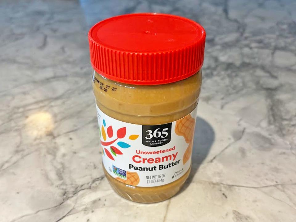 A jar of peanut butter with a white label with illustrations of peanuts and leaves  with a red lid