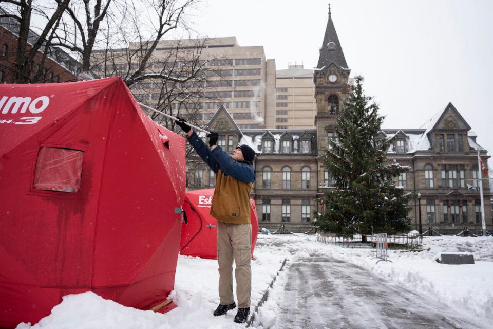 Matthew Grant uses a broom to clean snow off ice fishing enclosures at a tent encampment in front of City Hall in downtown Halifax, Monday, Dec. 4, 2023. The encampment was closed in March, and Grand Parade is expected to open this summer after repairs.