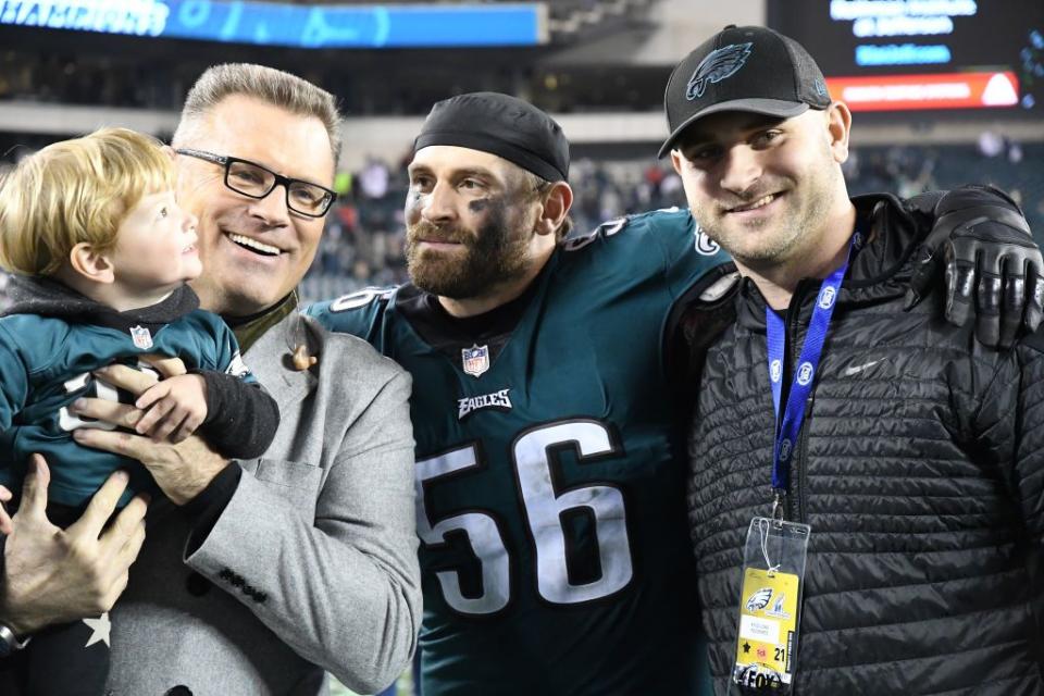 howie long smiles while holding a young child, chris long stands in a green football uniform on the right and smiles
