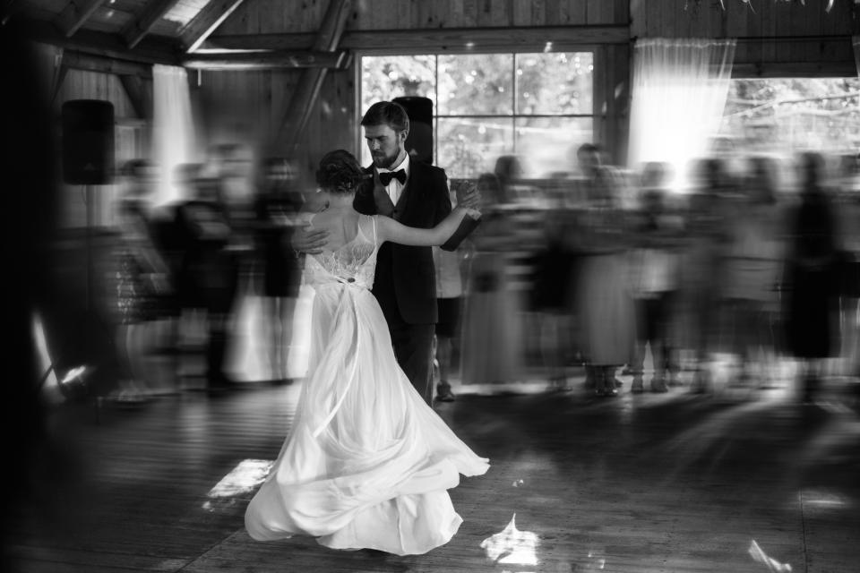 Bride and groom share their first dance in a rustic venue with blurred guests watching in the background. Bride wears a flowing gown, and groom is in a suit