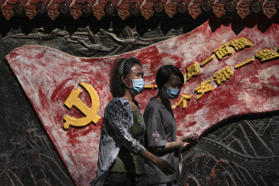 Women wearing face masks walk by a mural depicting a communist party flag and a party slogan in Beijing, Tuesday, Sept. 6, 2022. China has locked down millions of its citizens under tough COVID-19 restrictions and is discouraging domestic travel during upcoming national holidays. (AP Photo/Andy Wong)