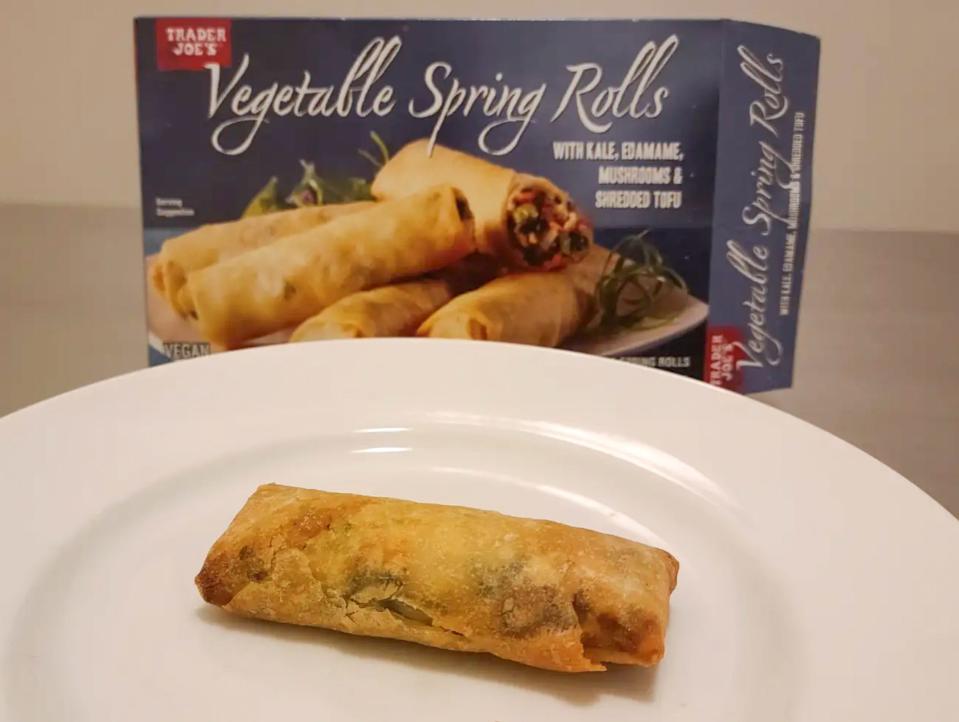 A golden-brown vegetable spring roll with a gray box of spring rolls behind the plate