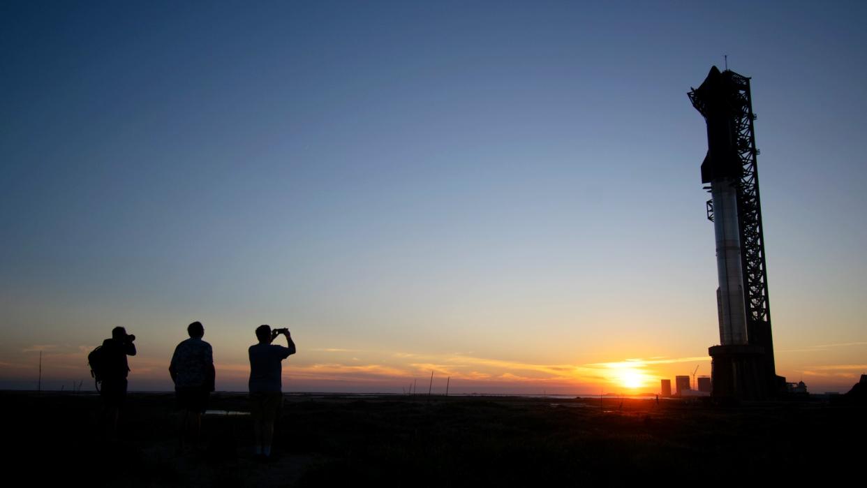  Silhouettes of three people at sunset in front of a large rocket on a launch pad in the background. 