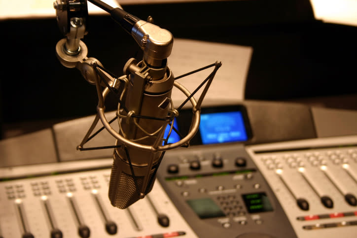 Studio microphone in focus with blurred mixing console in background, suggesting radio broadcast setting