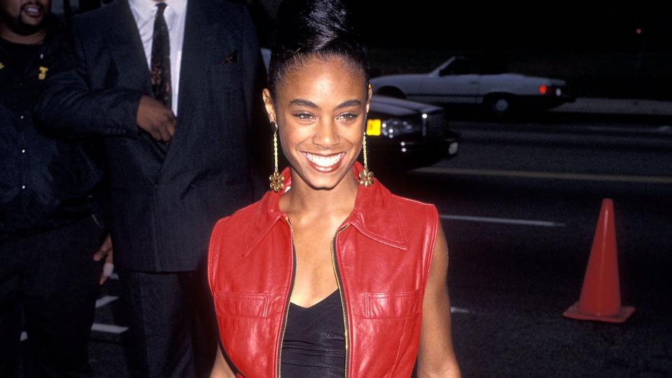jada pinkett smith wearing a red vest, black shirt and black pants, smiling for cameras while standing on a sidewalk outside