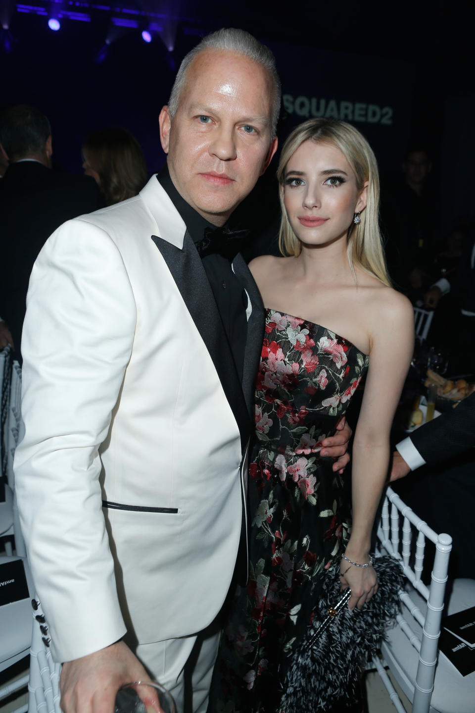 Ryan Murphy in a suit with black accents, poses with Emma Roberts wearing a floral strapless dress, at an event