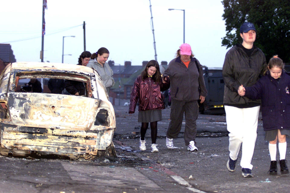 Local residents of the Glenbryn area in North Belfast walk past burnt-out remains of an armored police car following overnight rioting between Nationalists and Loyalists crowds January 10, 2002 in Belfast, Northern Ireland. Rioting erupted as Catholic parents picked up their children from the Holy Cross girls'' primary school.