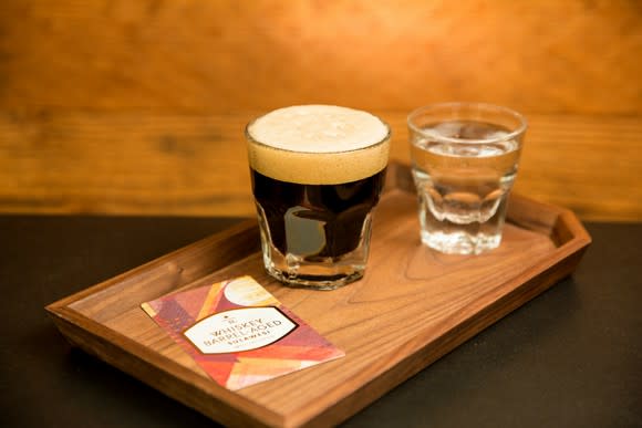 A glass of coffee is on a wooden board next to a glass of water.