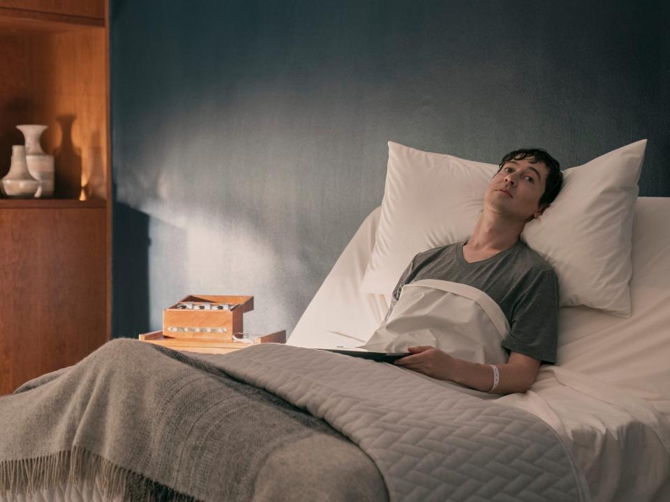 will downing laying in a comfortable bed in 3 body problem, holding a tablet connected to a container. the room is aesthetically pleasing, and saul is standing next to his bed