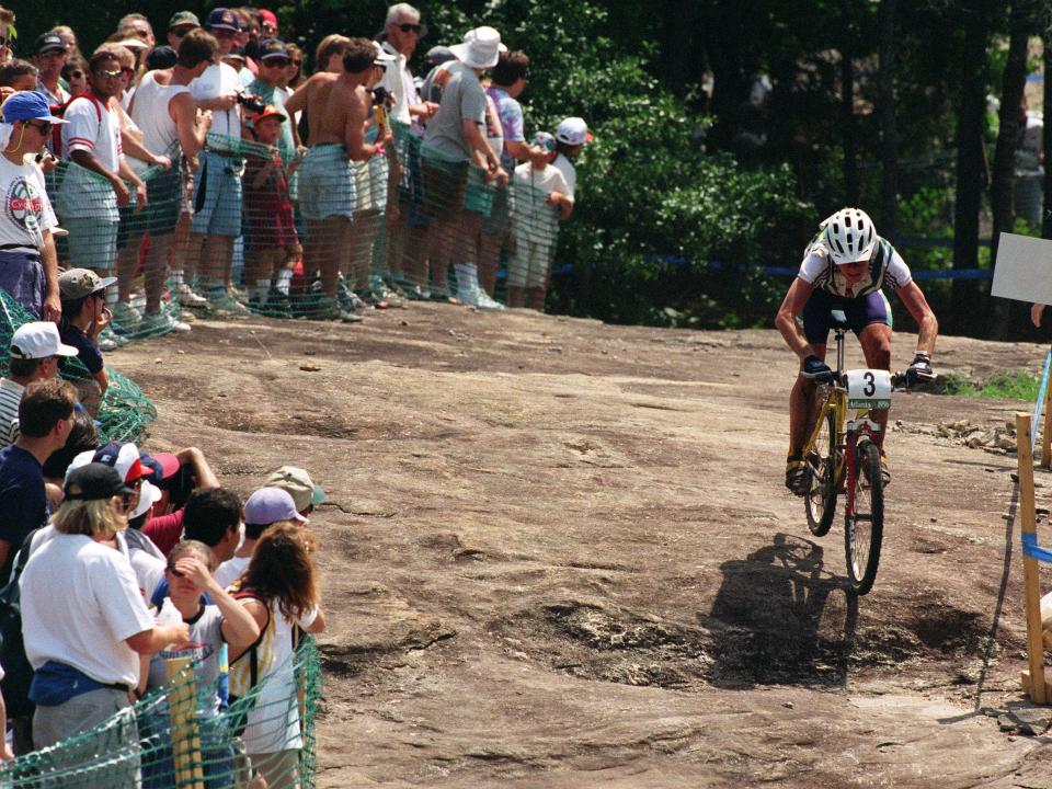 Mountain biker rides on the right while a crowd of people watch from behind a green fence on the left.