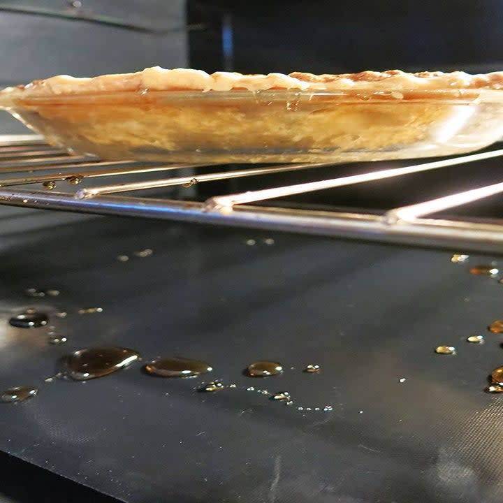 A pie baking in an oven lined at the bottom with one of the removable oven liners covered in drippings 