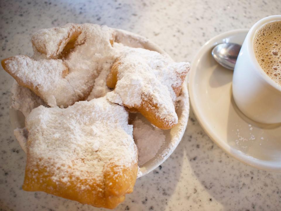 Beignets on with powered sugar on a plate beside a cup of coffee on a white table