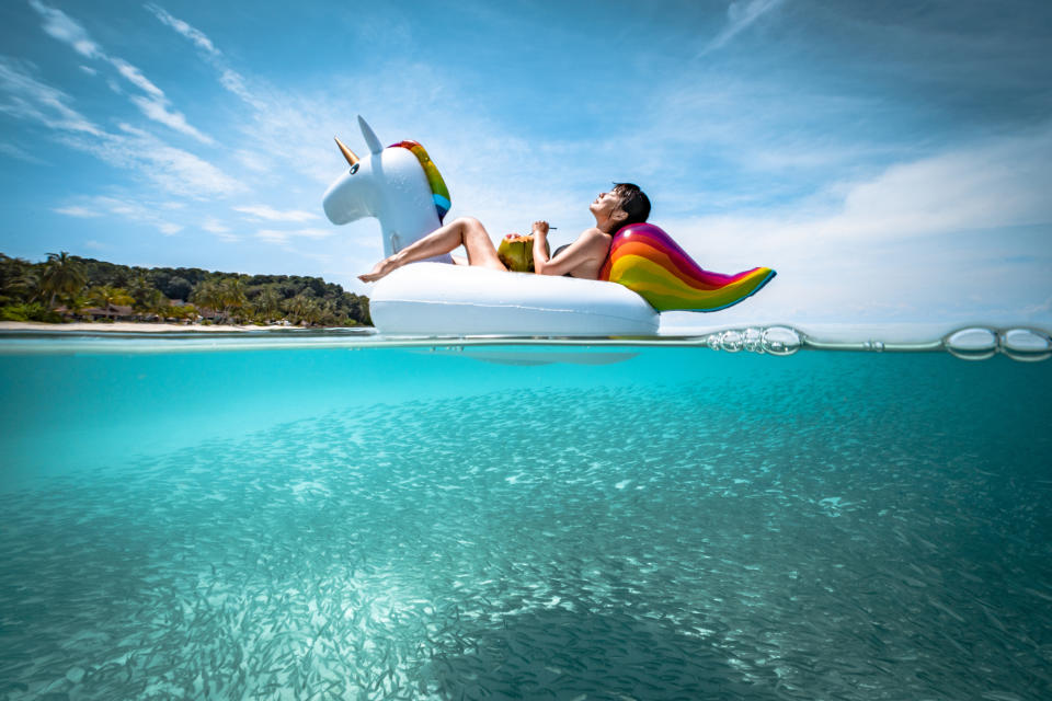 Girl holding a coconut enjoying the sun lying on a floating unicorn with fish swimming underwater. travel insurance