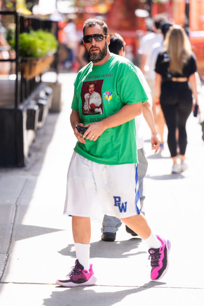 An unidentified man walks on the sidewalk wearing a green graphic t-shirt, white shorts with "PW" letters, and pink-accented sneakers