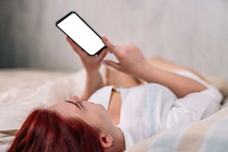 smiling young woman, looking at her social networks on cell phone, lying on her bedroom bed.