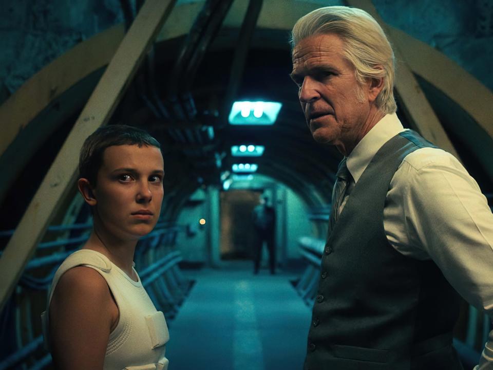 millie bobby brown as eleven and matthew modine as dr. brenner in stranger things, standing together in a long, underground hallway, while looking not at each other, but towards the camera