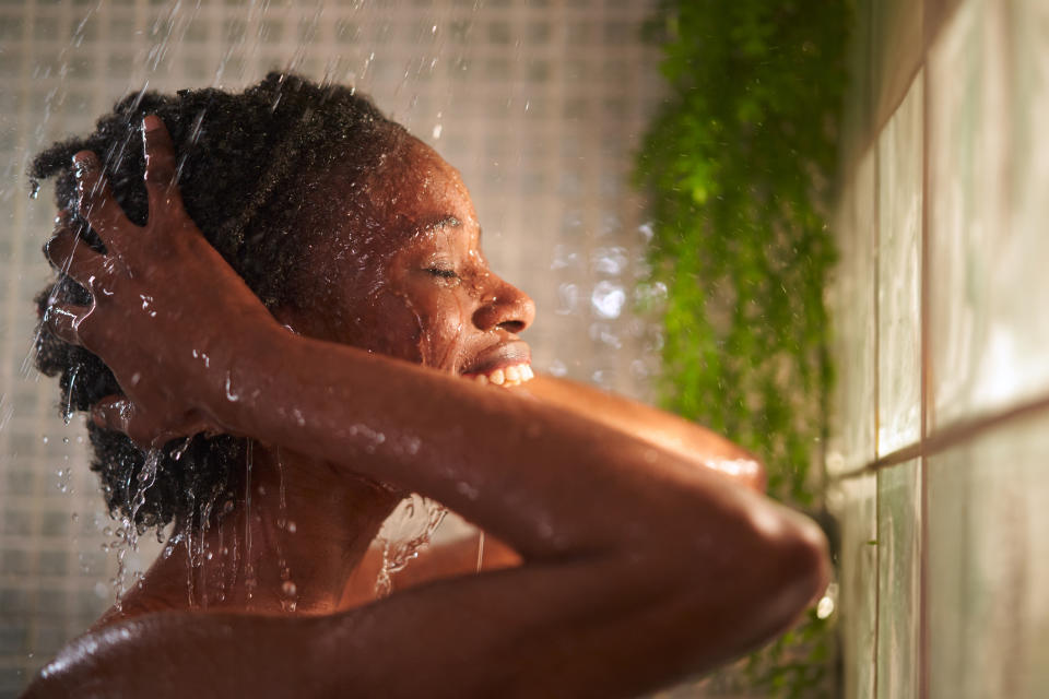 A person with a joyful expression showers, water streaming over their head and shoulders, with green plants seen on the tiled wall in the background