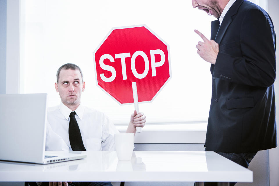 A business man sits at his office desk in front of his computer, while his CEO yells loudly at him.  He holds up a stop sign with a funny look on his face, ready to be finished with his job.  Horizontal.