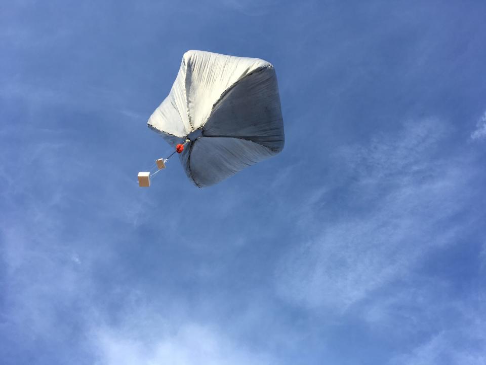 pentagon shaped white balloon with boxes dangling from a string below rises into a blue sky with whisps of cloud