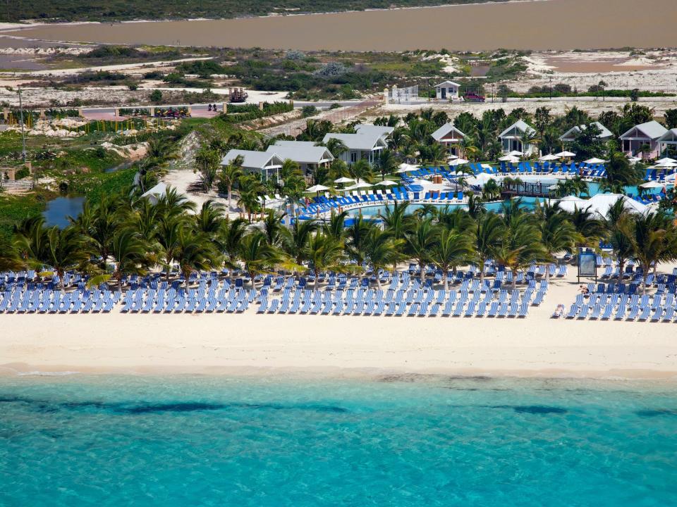 rows of lounge chairs on the beach in front of a blue ocean. Behind the chairs, there's rows of palm trees, a pool, and small buildings