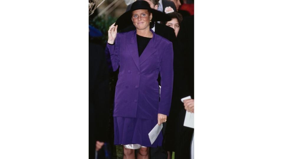 Sarah, Duchess of York wearing a baby bump-skimming purple outfit and holding her hat at the wedding of the Marquis of Marlborough and Rebecca Few Brown