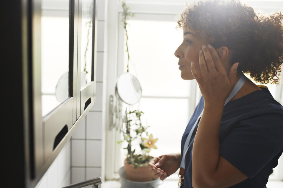 A woman with curly hair, wearing a lanyard and scrubs, checks her reflection in a bathroom mirror, adjusting her earring