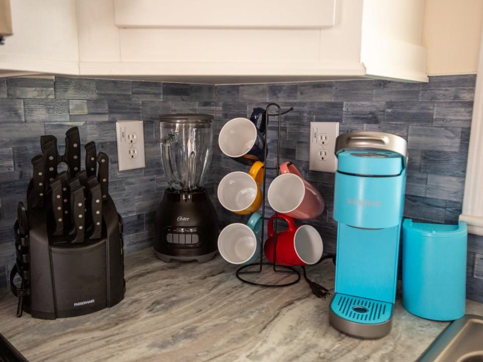 A kitchen with a coffee machine, mugs on a rack, knives, and a blender.