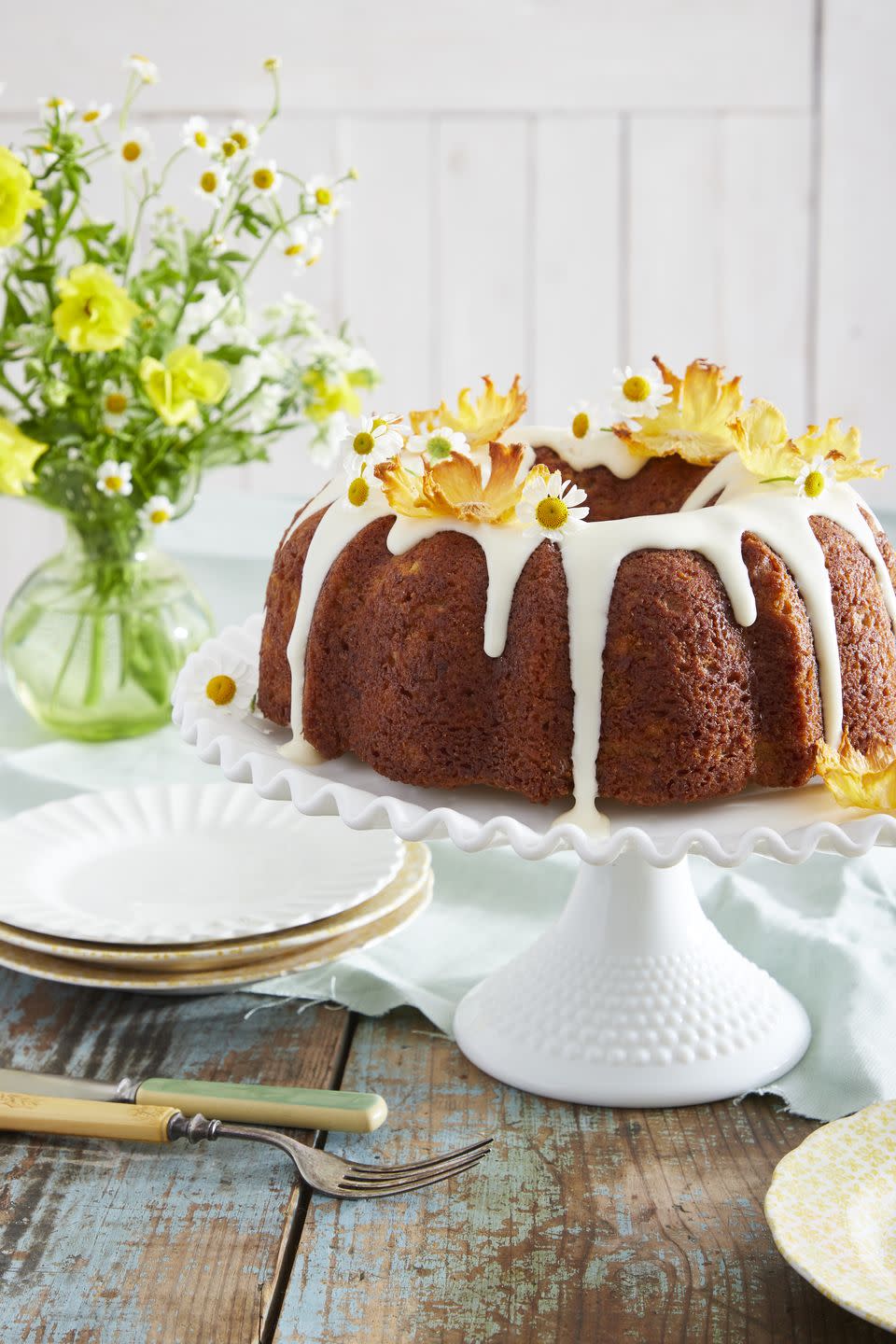 hummingbird bundt cake on a white cake stand and topped with cream cheese glaze and garnished with dried pineapple flowers and fresh camomile flowers