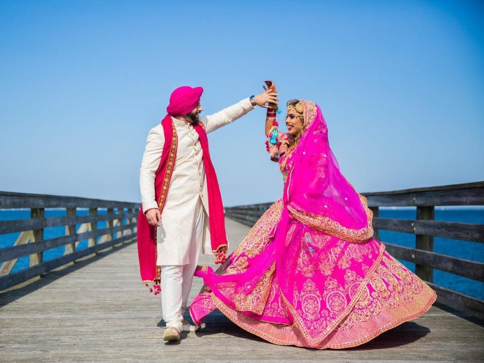 A groom and bride dance on a boardwalk wearing traditional Indian wedding attire.