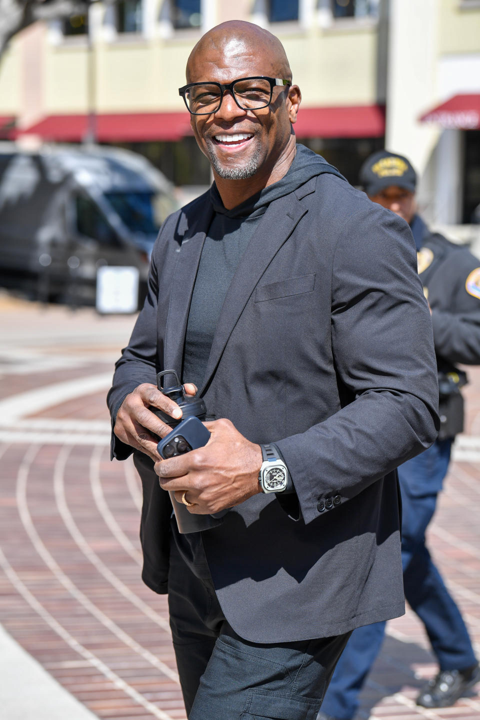 Terry Crews smiles while walking outside, wearing a black blazer over a black shirt and holding his phone and glasses case