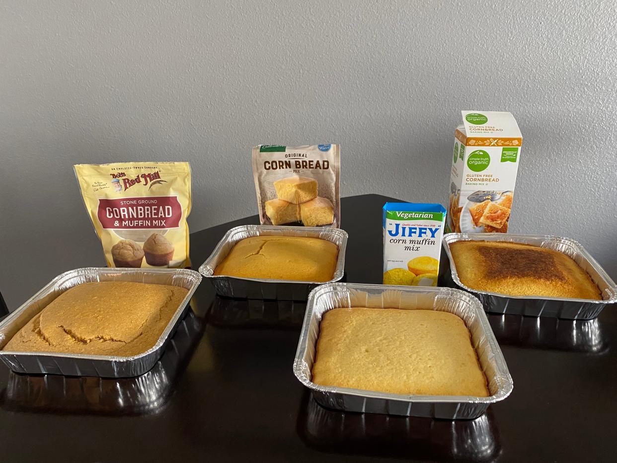 Four cornbread packages from Bob's Red Mill, Marie Callender's, Jiffy, and Simple Truth Organic on black table beside cooked trays