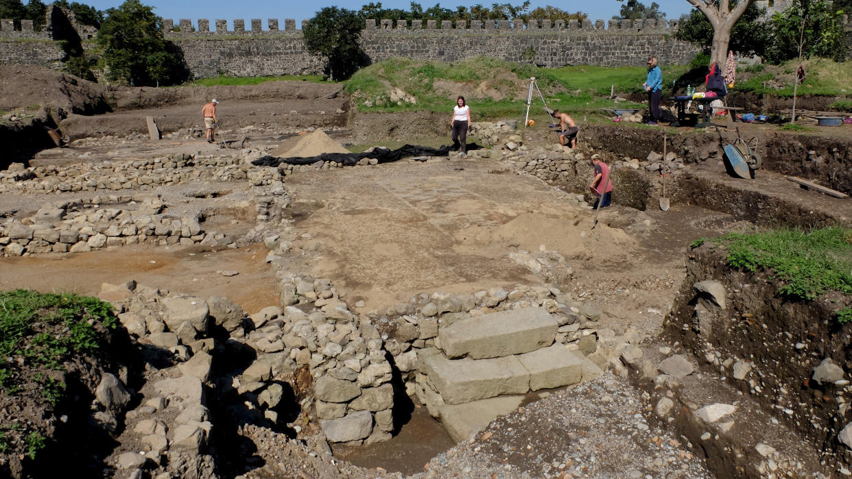  We see an archaeological site with different levels of dirt and stone. There are five people excavating at the site, which has a stone wall in the background. 