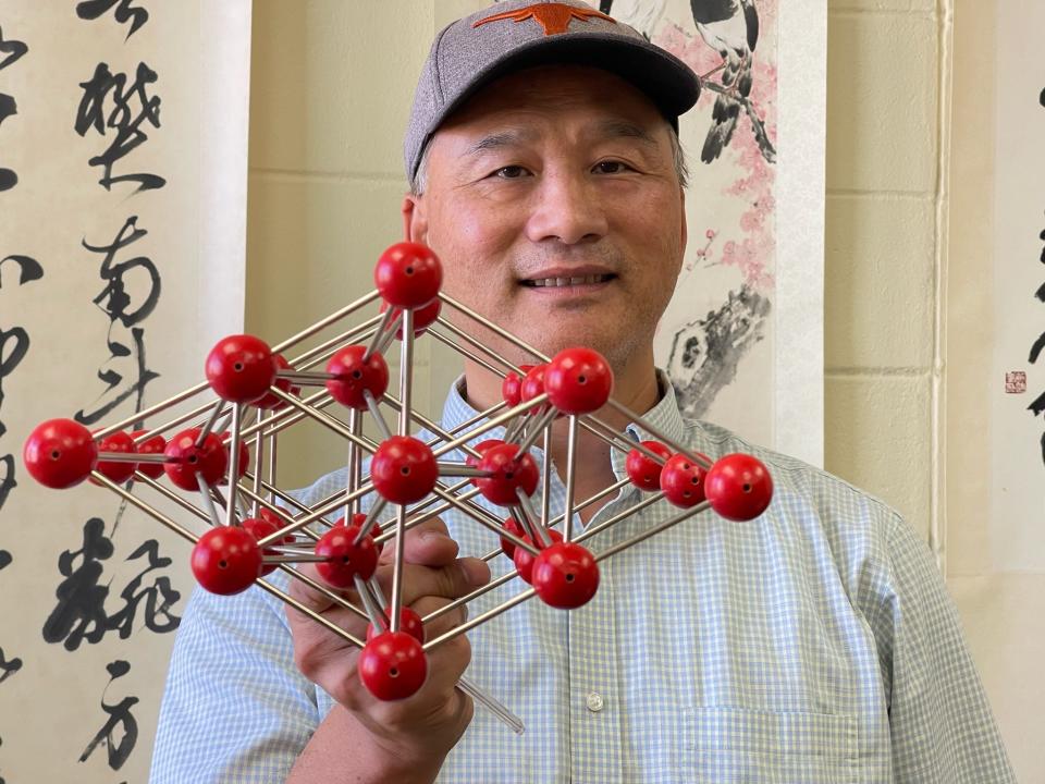 A scientist holds up a structure made of red balls and metal rods, representing atoms in a hexagonal pattern the inner core. The scientist, wearing a cap adorned with a bull's head and smiling at the camera, is standing in front of wall art. 