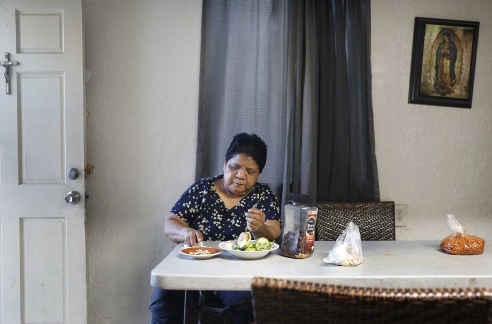 A woman sits at a kitchen table with plates of food in front of her. A picture of the Virgin of Guadalupe hangs on the wall.