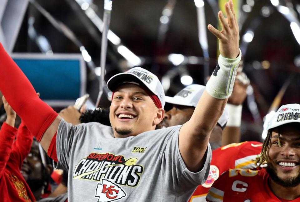 patrick mahomes smiles and raises his arms in celebration, wearing a grey superbowl champions shirt and similar light grey hat, people stand behind and around him