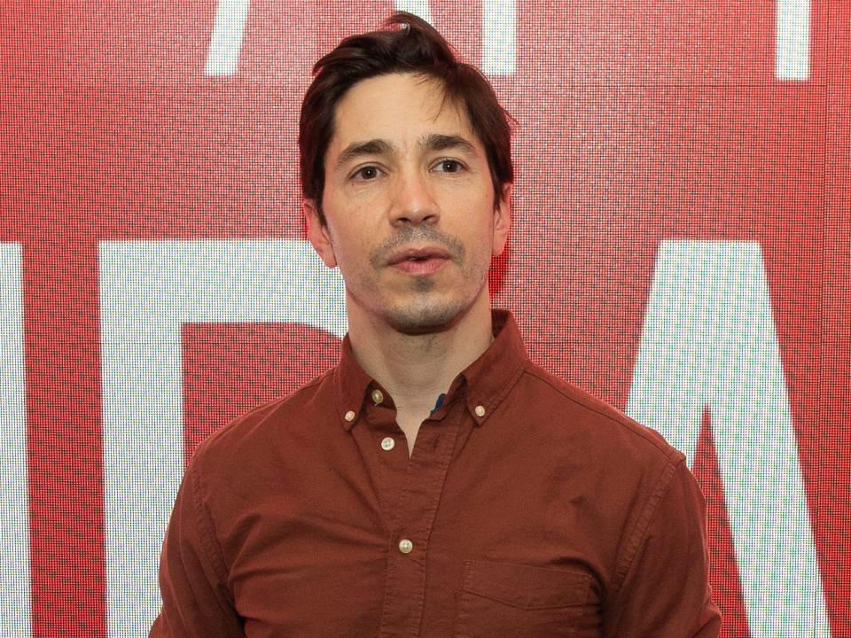 Justin Long in an orange button-down shirt in front of a red background