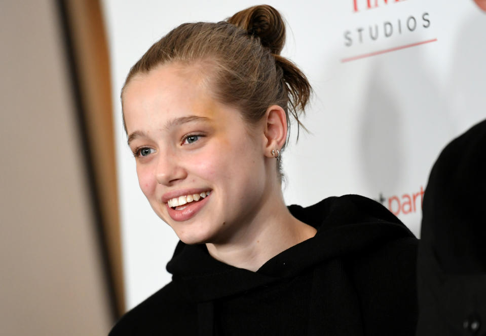 Shiloh Jolie-Pitt, wearing a casual black hoodie, smiles at a red carpet event