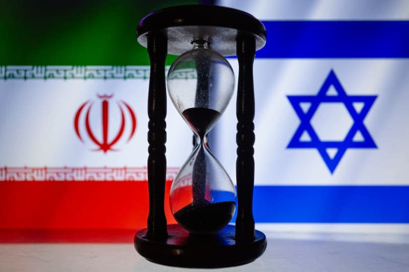 Iran and Israel cropped flags are displayed behind a wooden hourglass sand timer. Andre M. Chang/ZUMA Press Wire/dpa