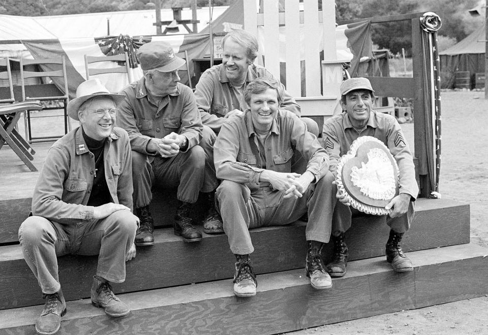 FILE - Cast members of the television series "M*A*S*H" take a break on the set during taping in Los Angeles on Sept. 15, 1982. The actors, from left, are, William Christopher, Harry Morgan, Mike Farrell, Alan Alda, and Jamie Farr. The combat boots and dog tags that Alda wore every day as he portrayed the wisecracking surgeon Hawkeye on the beloved television series "M*A*S*H" meant so much to him that when the show ended 40 years ago, he took them with him. But now, he's ready to let them go to benefit another passion: his center that helps scientists communicate better. Heritage Auctions is offering up the worn boots and dog tags on July 28 in Dallas. (AP Photo/Wally Fong, File)