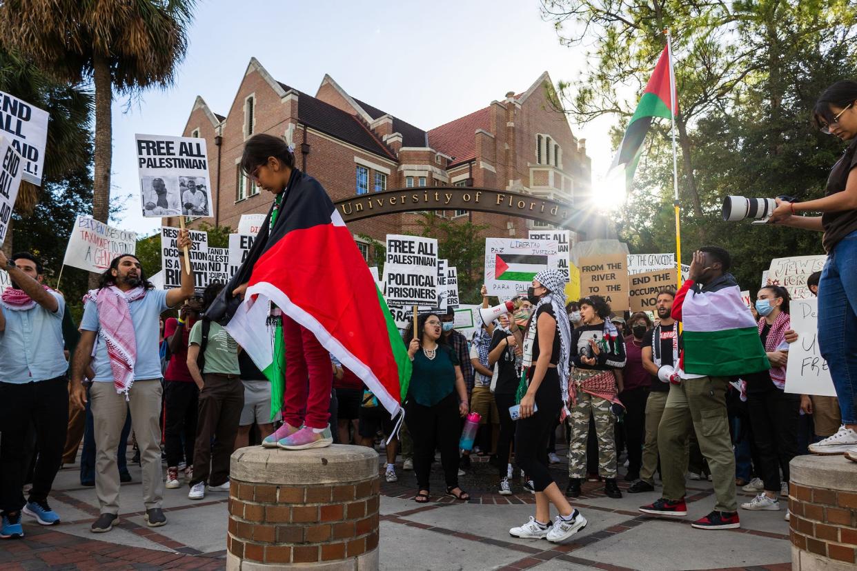 A girl wrapped in a Palestinian flag stands on a bollard as protestors advocating for the end of the siege on Gaza gather on Oct. 25, 2023, on the corner of University Avenue and 13th Street at the University of Florida in Gainesville.