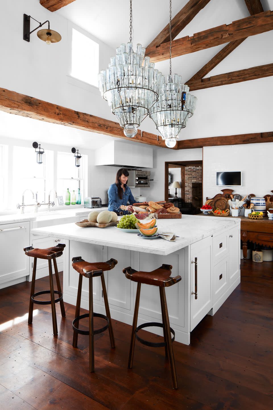 an all white kitchen with wood beams and a large island in the middle where a woman is standing